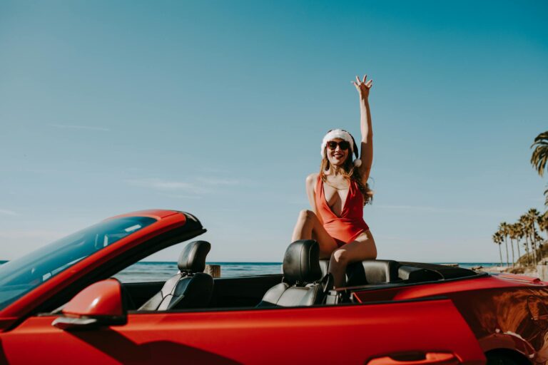 A woman in a Santa hat enjoying a festive moment in a red convertible by the seaside.